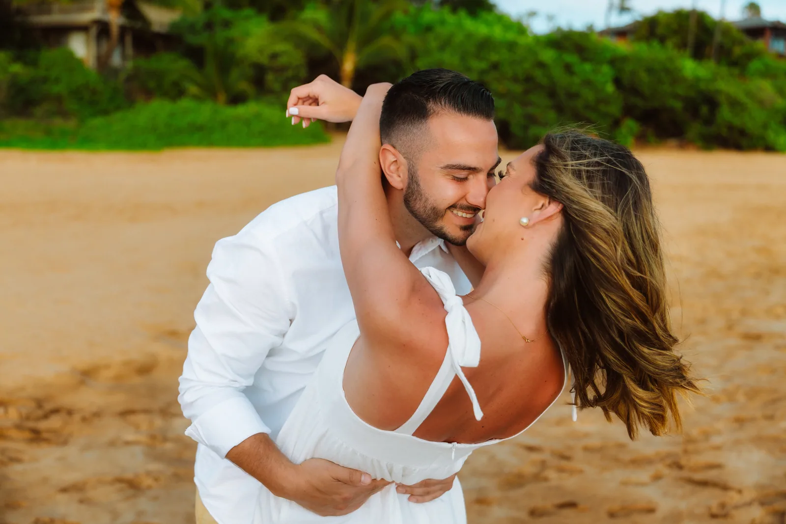 A couple laughing mid-dip-kiss on the sand below a green hillside