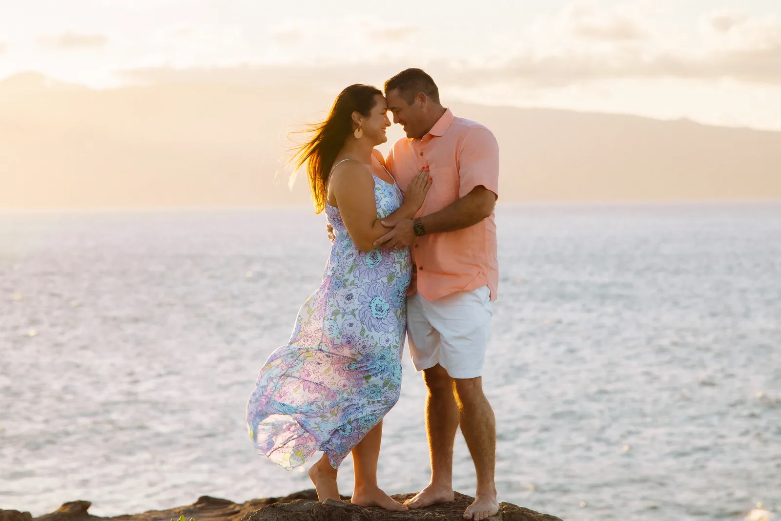 A couple standing close on a sea cliff as the wind lifts her dress