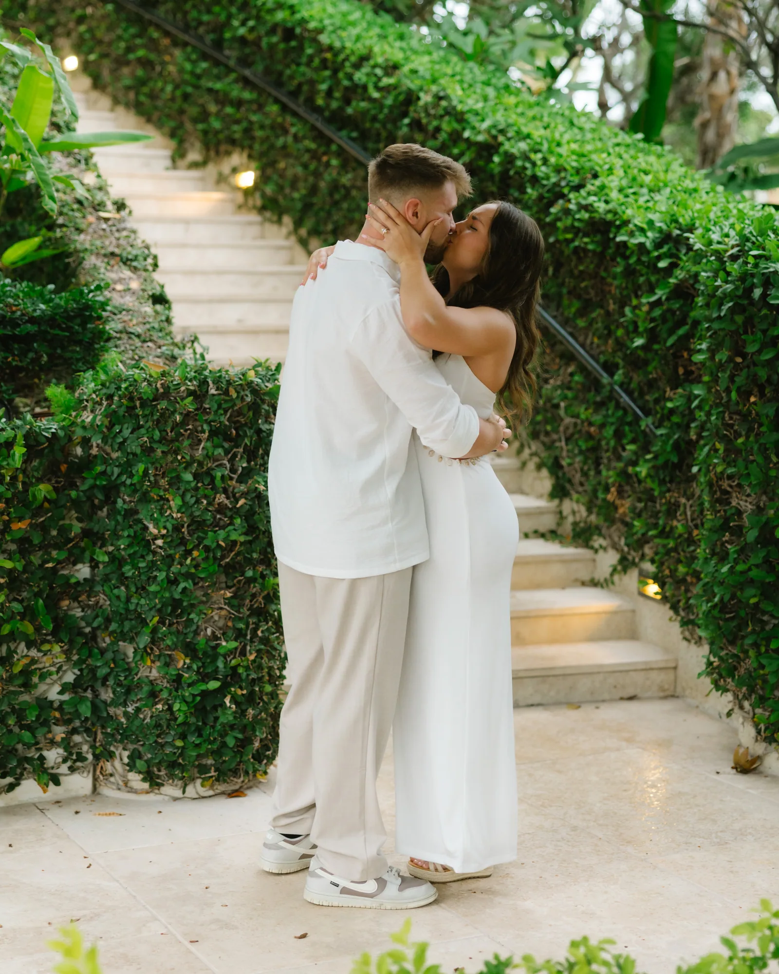 A couple in white kissing on a hedged garden staircase