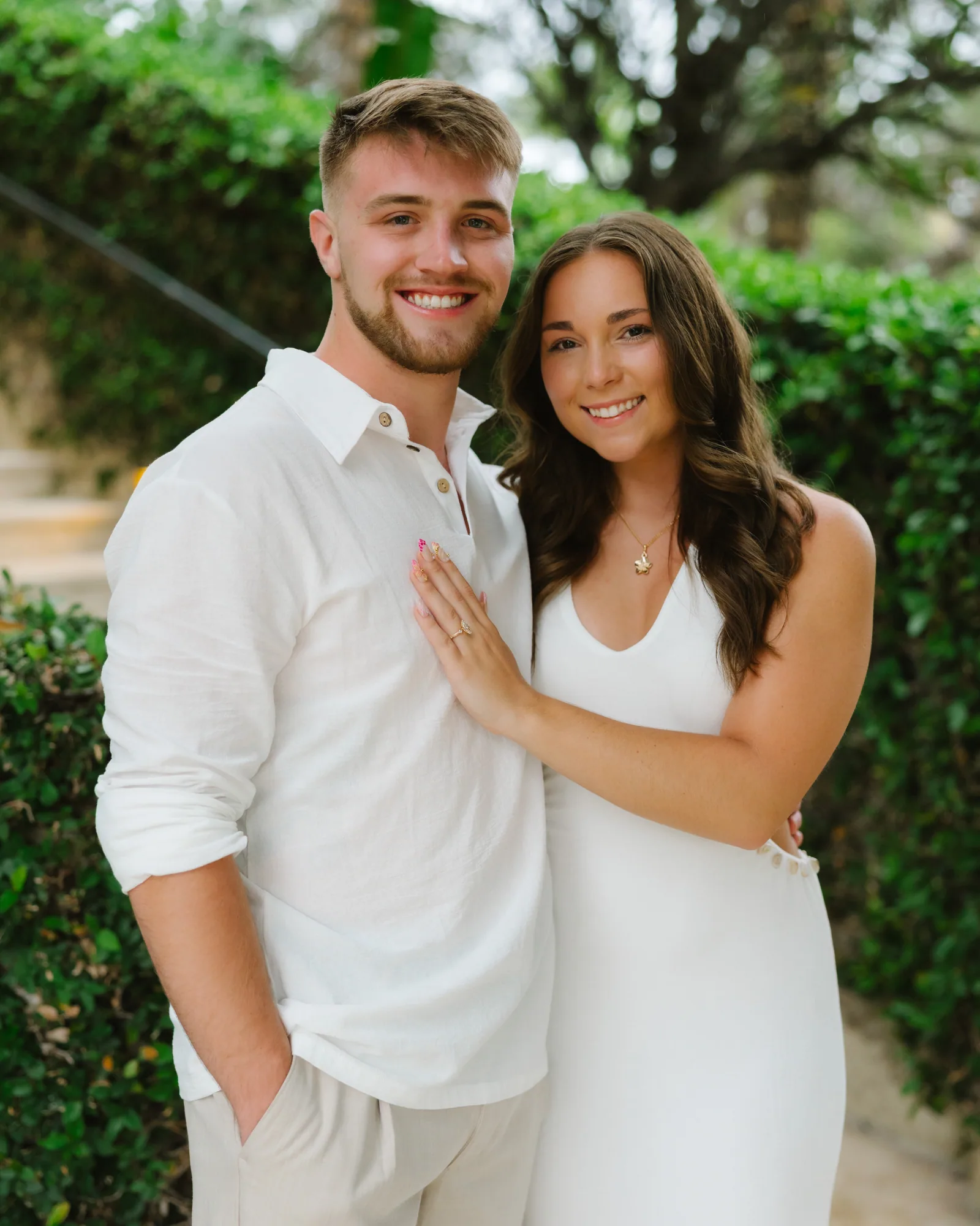 A couple in white photographed together against a garden hedge