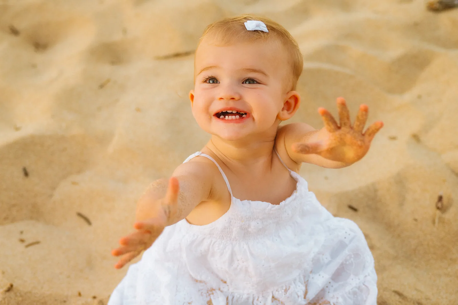 A baby sitting in the sand reaching toward the camera, mid-laugh