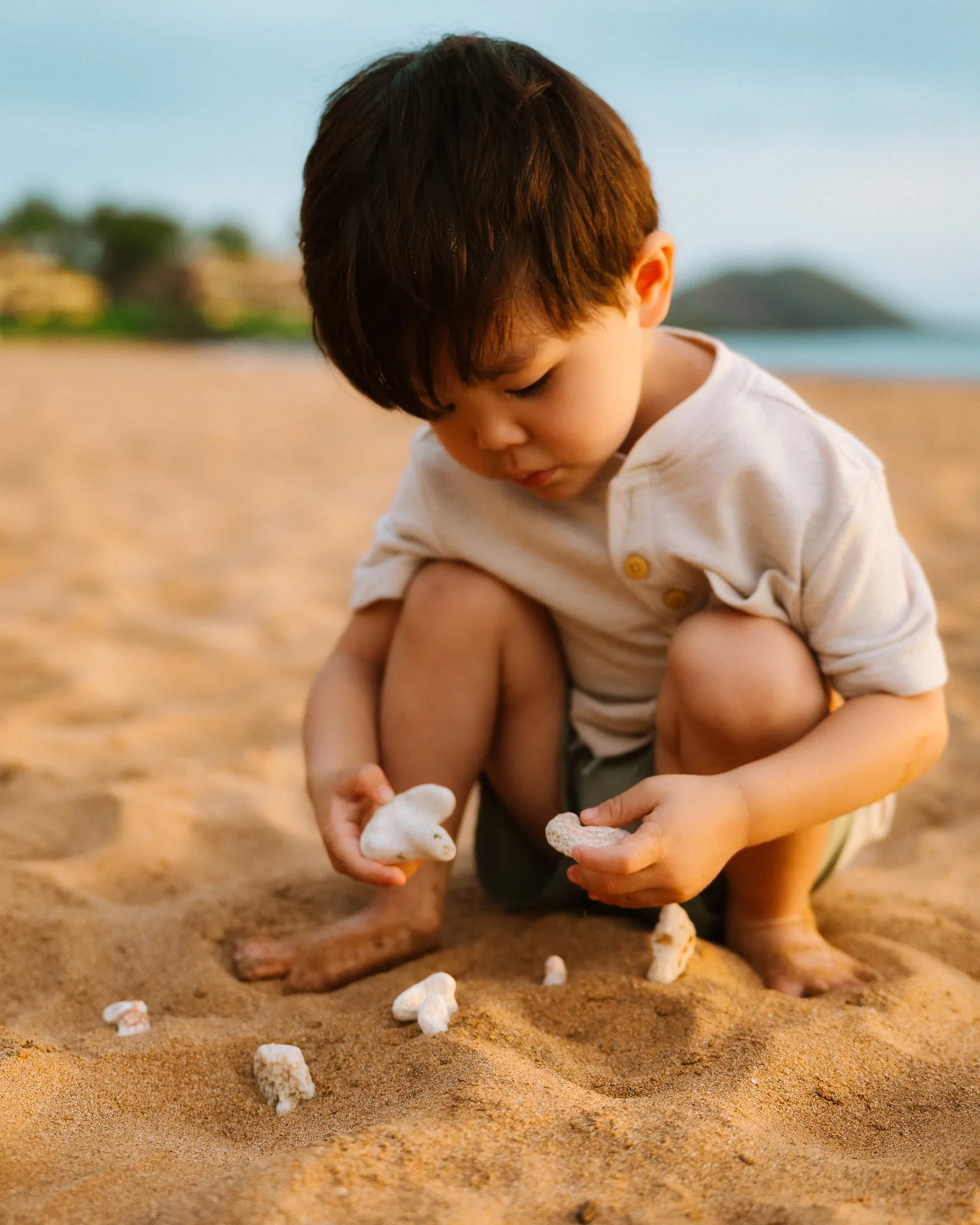 A young boy crouched in the sand studying the shells in his hands
