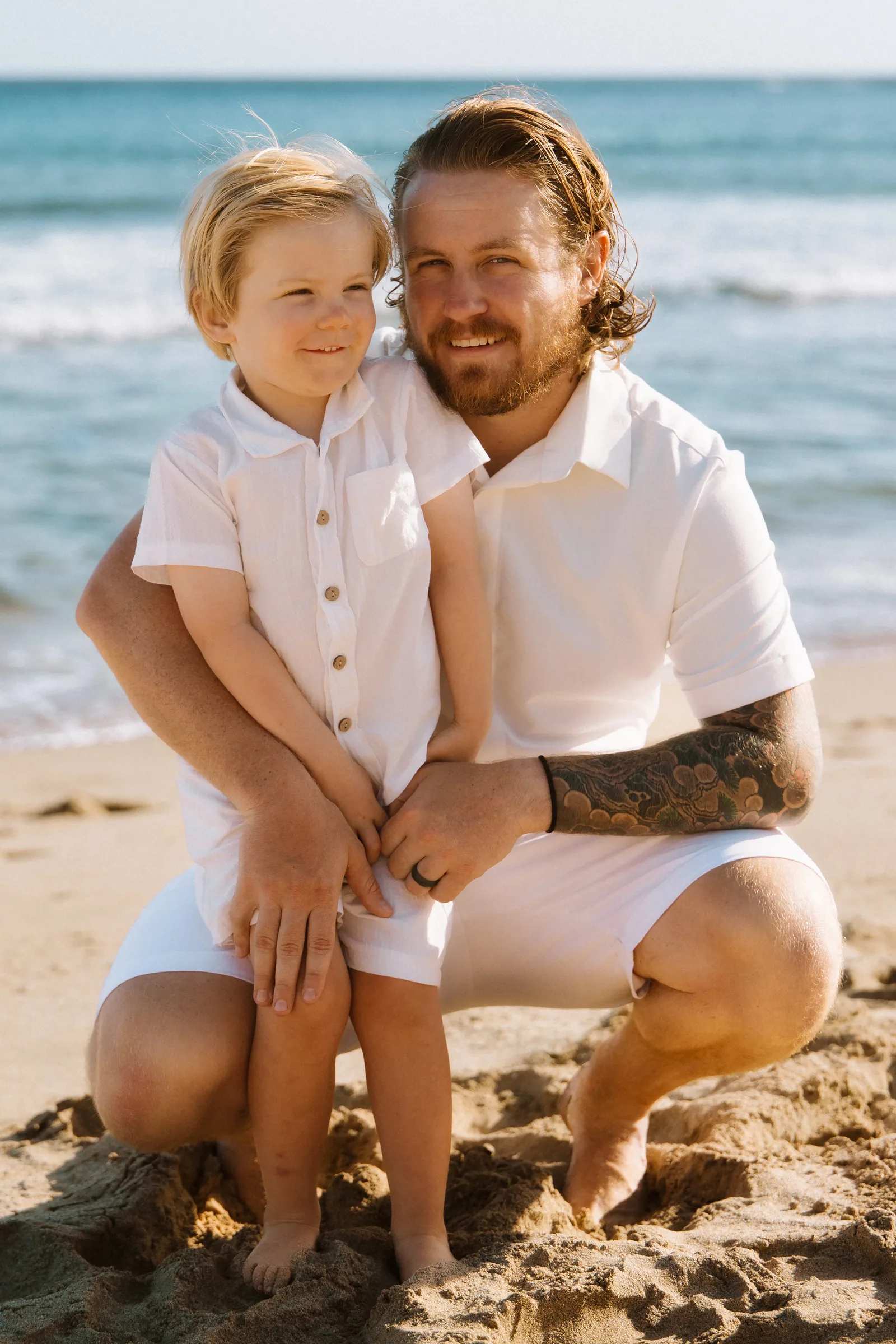 A father crouched beside his young son on the sand, both in white