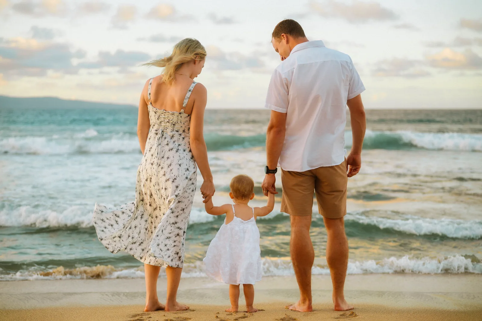 Parents and their toddler standing at the water's edge, seen from behind
