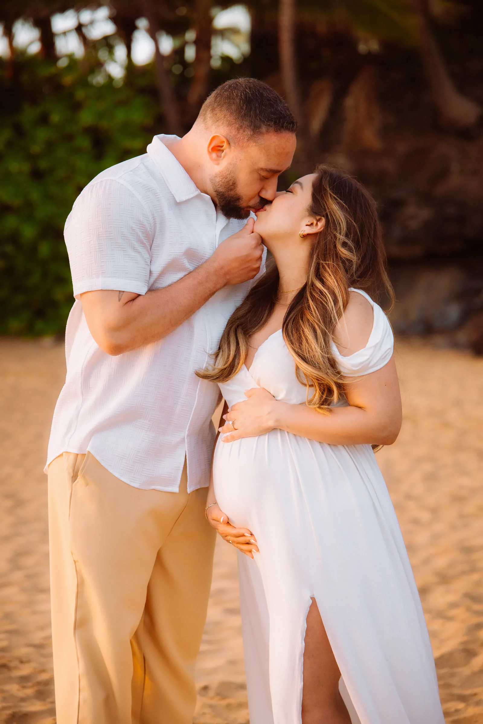 An expectant couple kissing on the sand in white