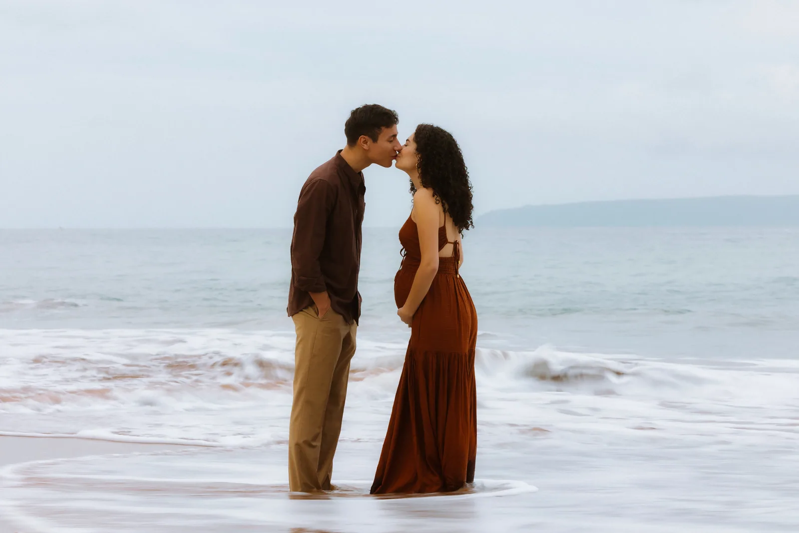 An expectant couple kissing in the shallows at dusk