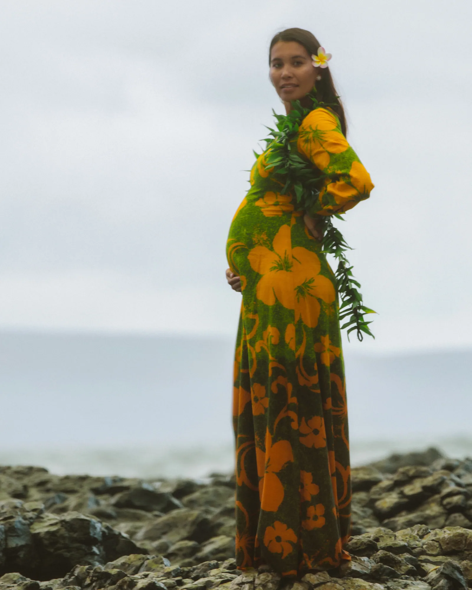 An expectant mother in a floral gown and lei among the rocks