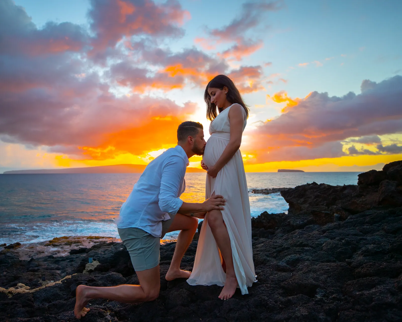 A partner kneeling to kiss the bump as the sun goes down