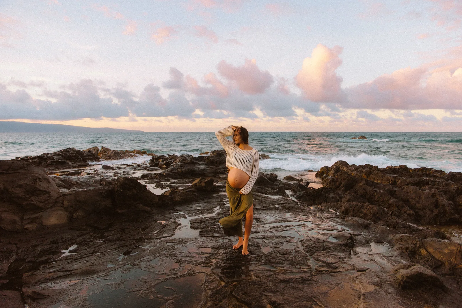 An expectant mother standing on lava rock under a pastel sky