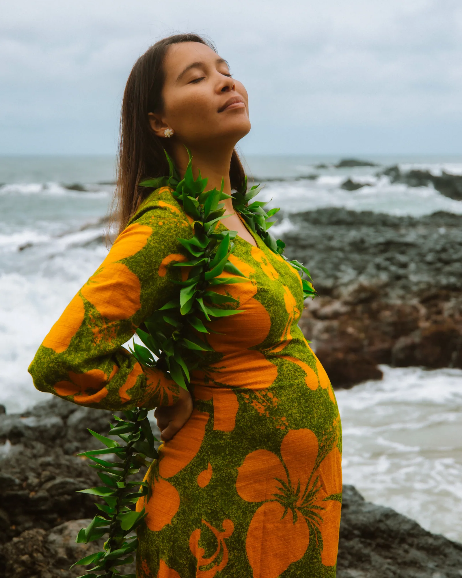 An expectant mother in a lei with her eyes closed, coastline behind