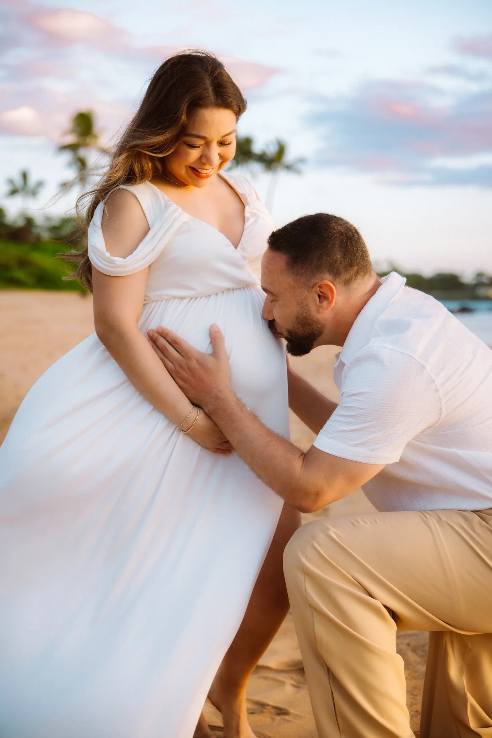 A partner leaning in to kiss the bump, white gown on the sand