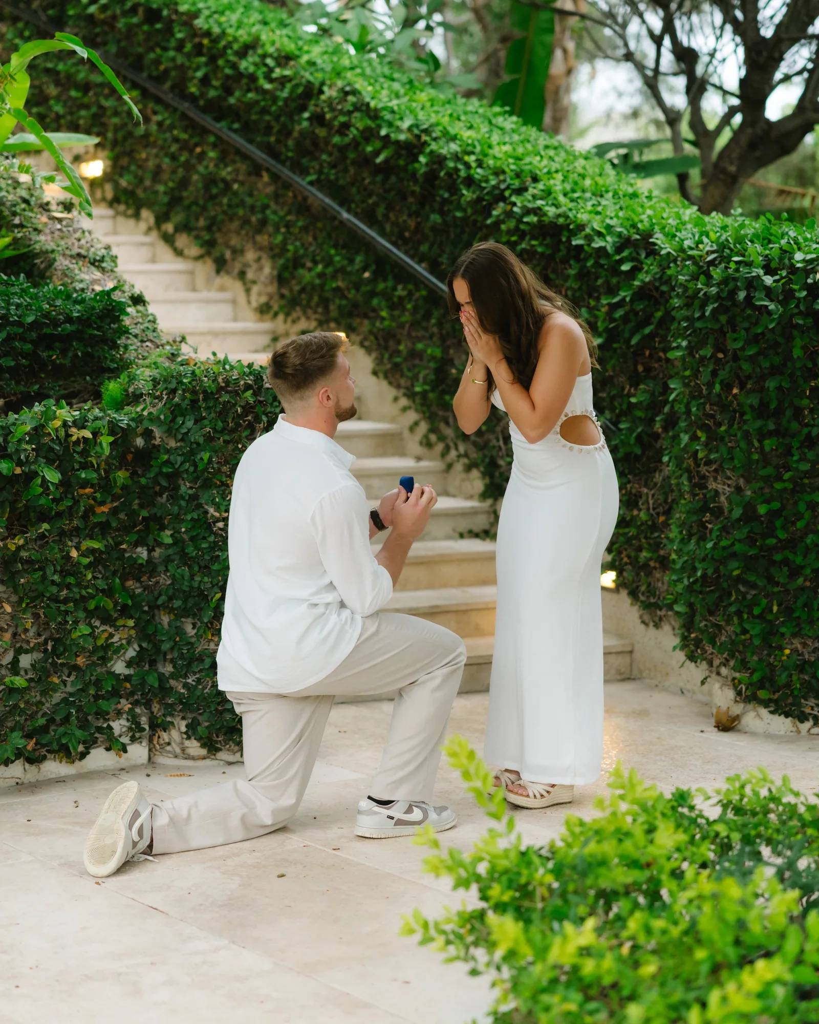 A proposal on lit garden stairs, him kneeling with the ring box open