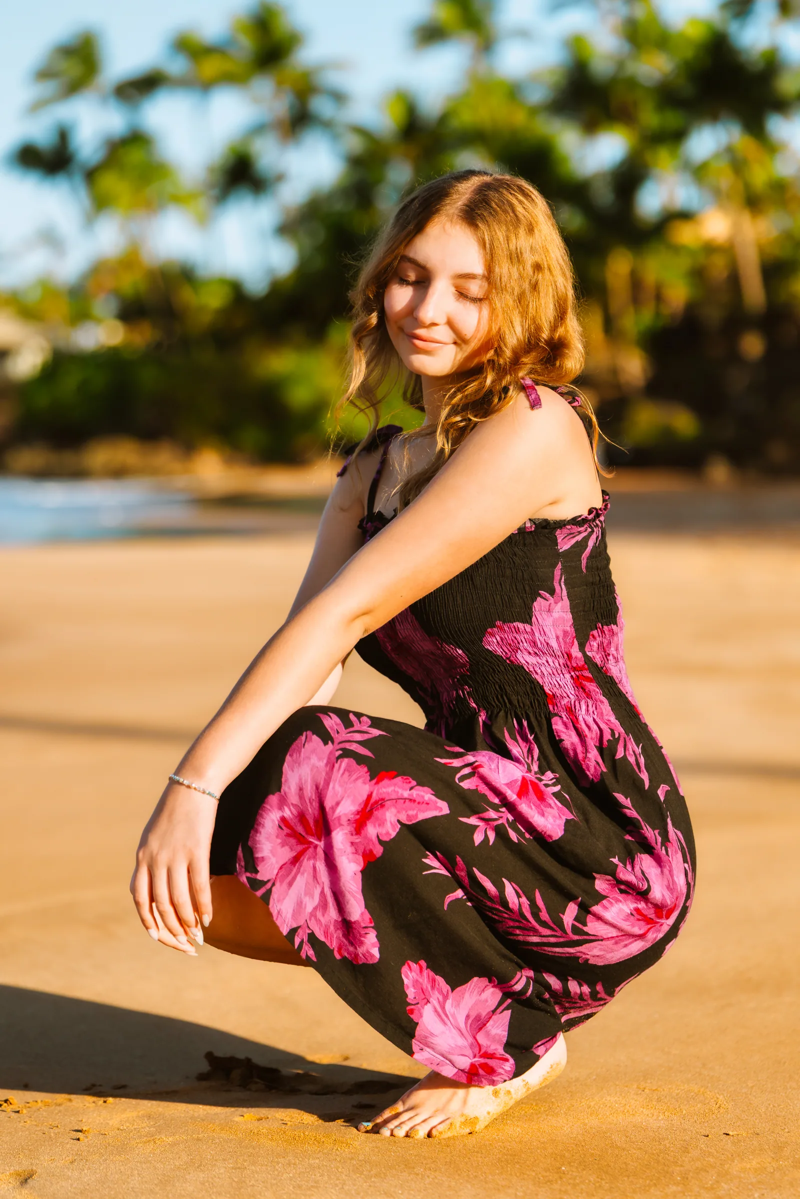 A senior portrait crouched on the sand in a floral dress