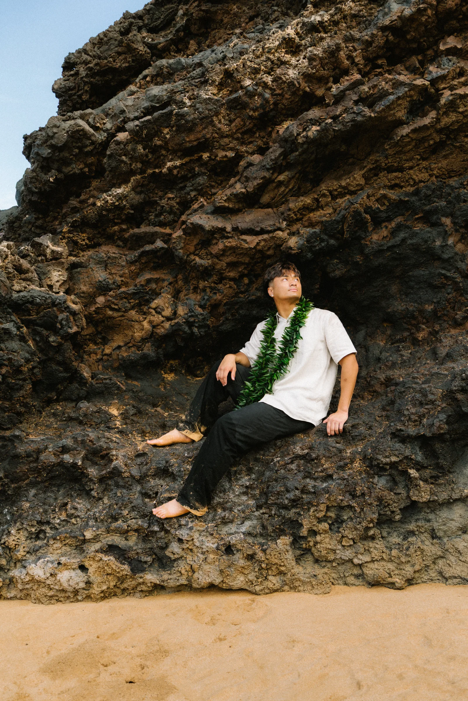 A senior portrait seated against lava rock, wearing a graduation lei