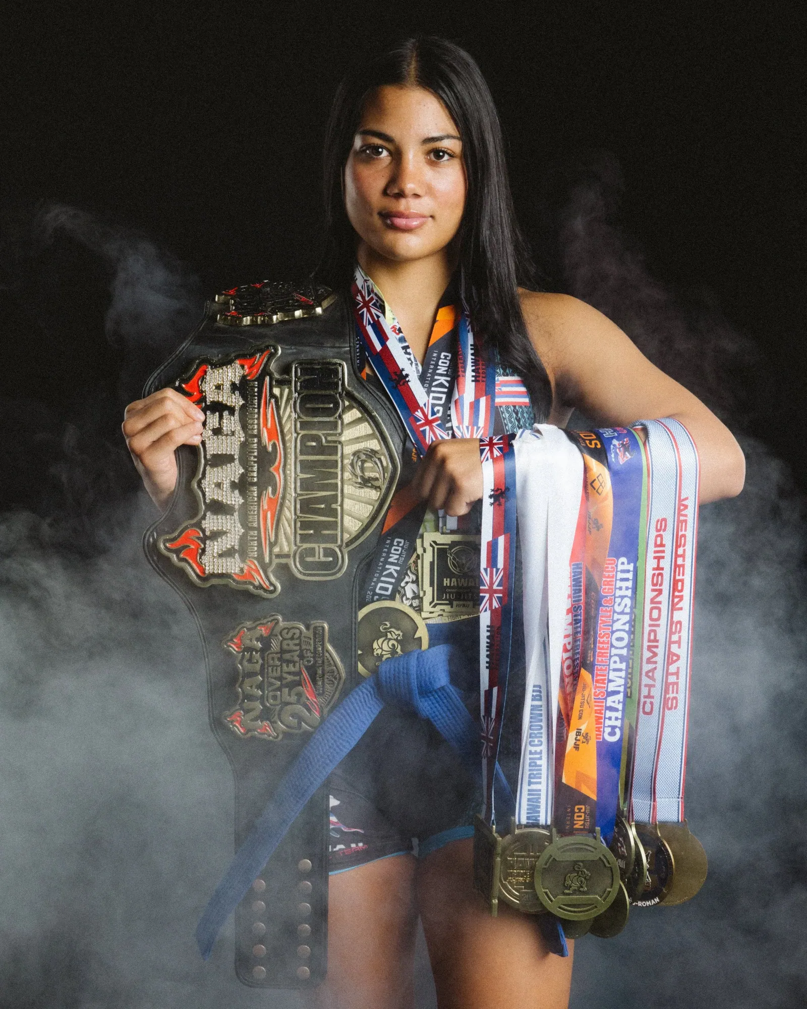 Jiu-jitsu champion holding her title belt and medals in studio smoke against a black backdrop