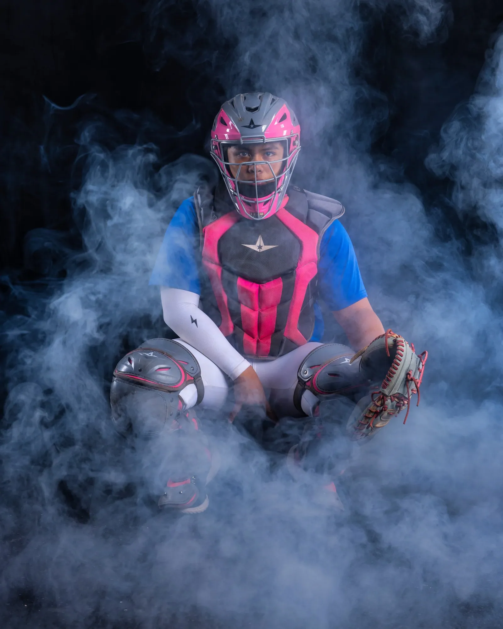 Softball catcher in full gear seated in studio smoke against black