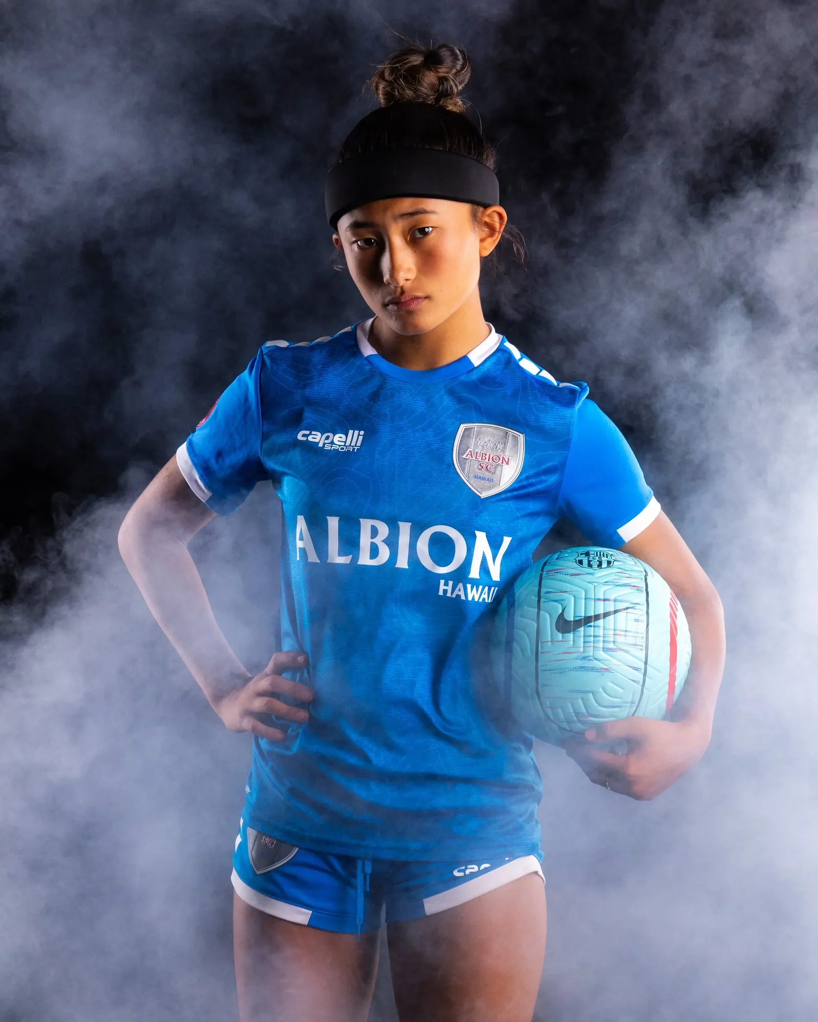 Soccer player standing with the ball on her hip, studio smoke against black